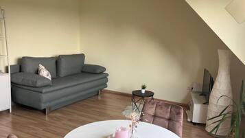 Ein modernes Wohnzimmer mit einem grauen Sofa, einem runden Esstisch mit Stühlen und einem kleinen Beistelltisch mit einer Pflanze.