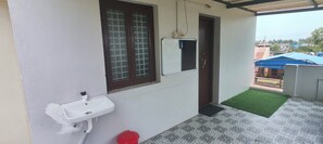 Une petite construction d’une seule pièce, à l’extérieur blanc, avec une fenêtre à cadre marron, un lavabo blanc et une porte donnant sur un espace extérieur doté d’un tapis vert.