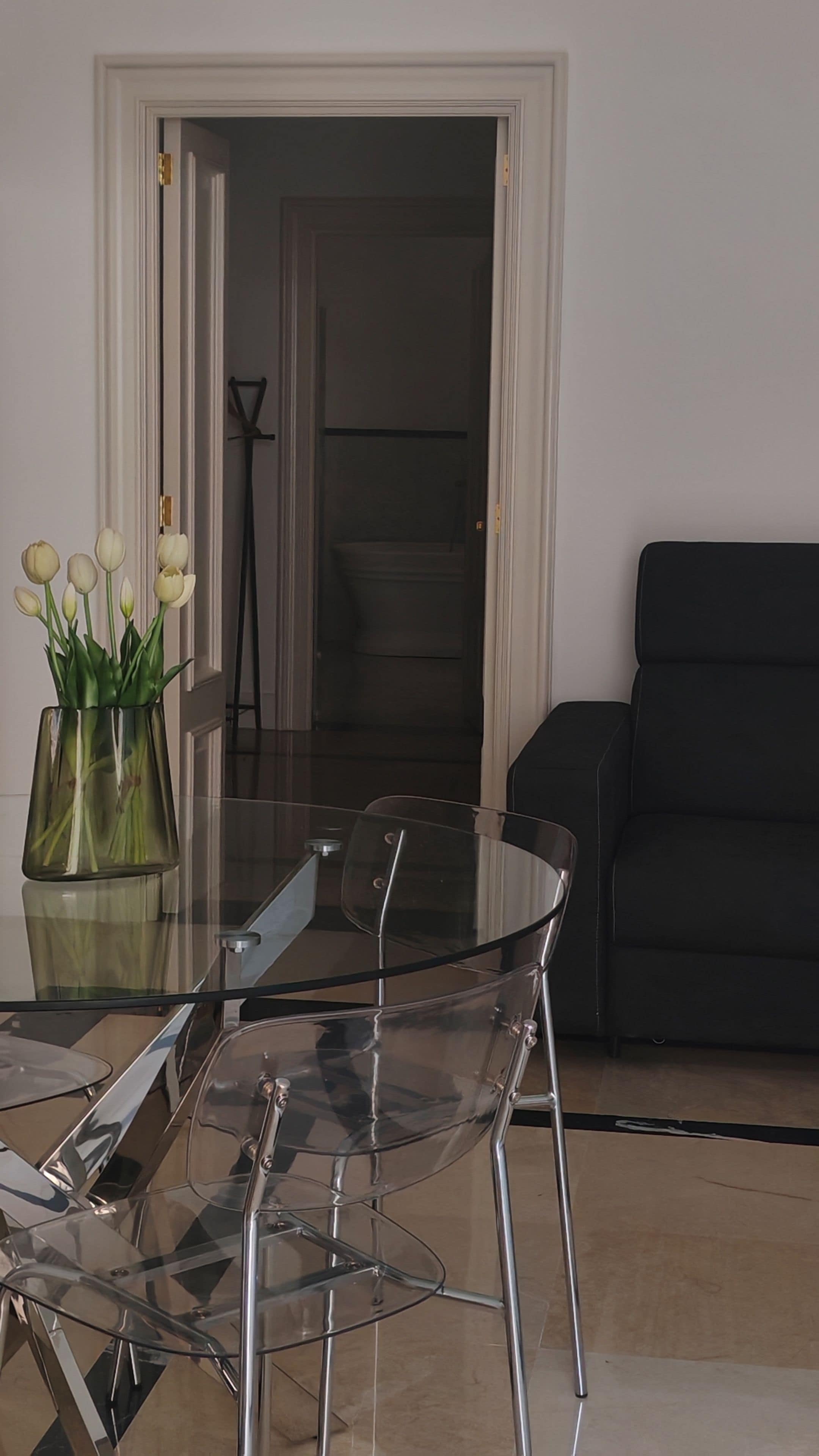 A dining area with a glass table and chairs, a vase with flowers, and a black sofa in the background.