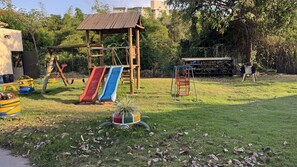 Un parque infantil con toboganes y columpios, rodeado de vegetación y una zona vallada.