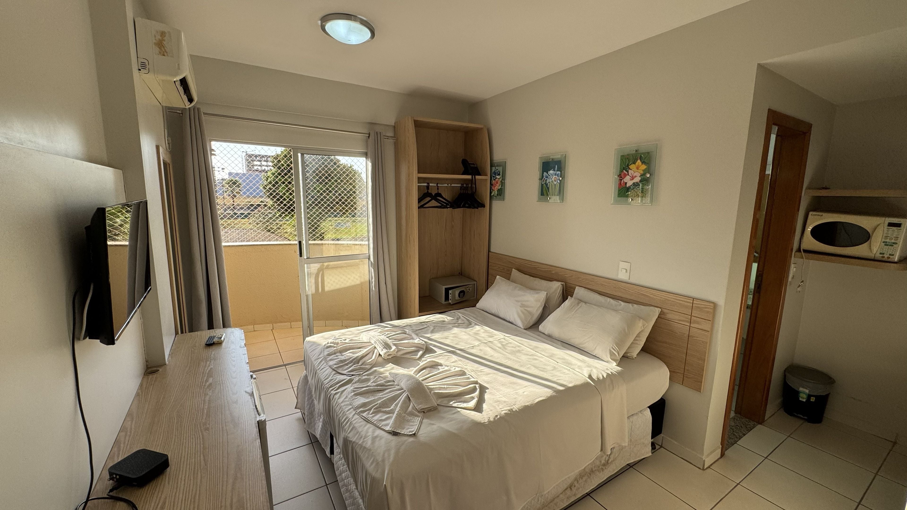 Quarto de hotel com cama, escrivaninha, televisão, armário e varanda.