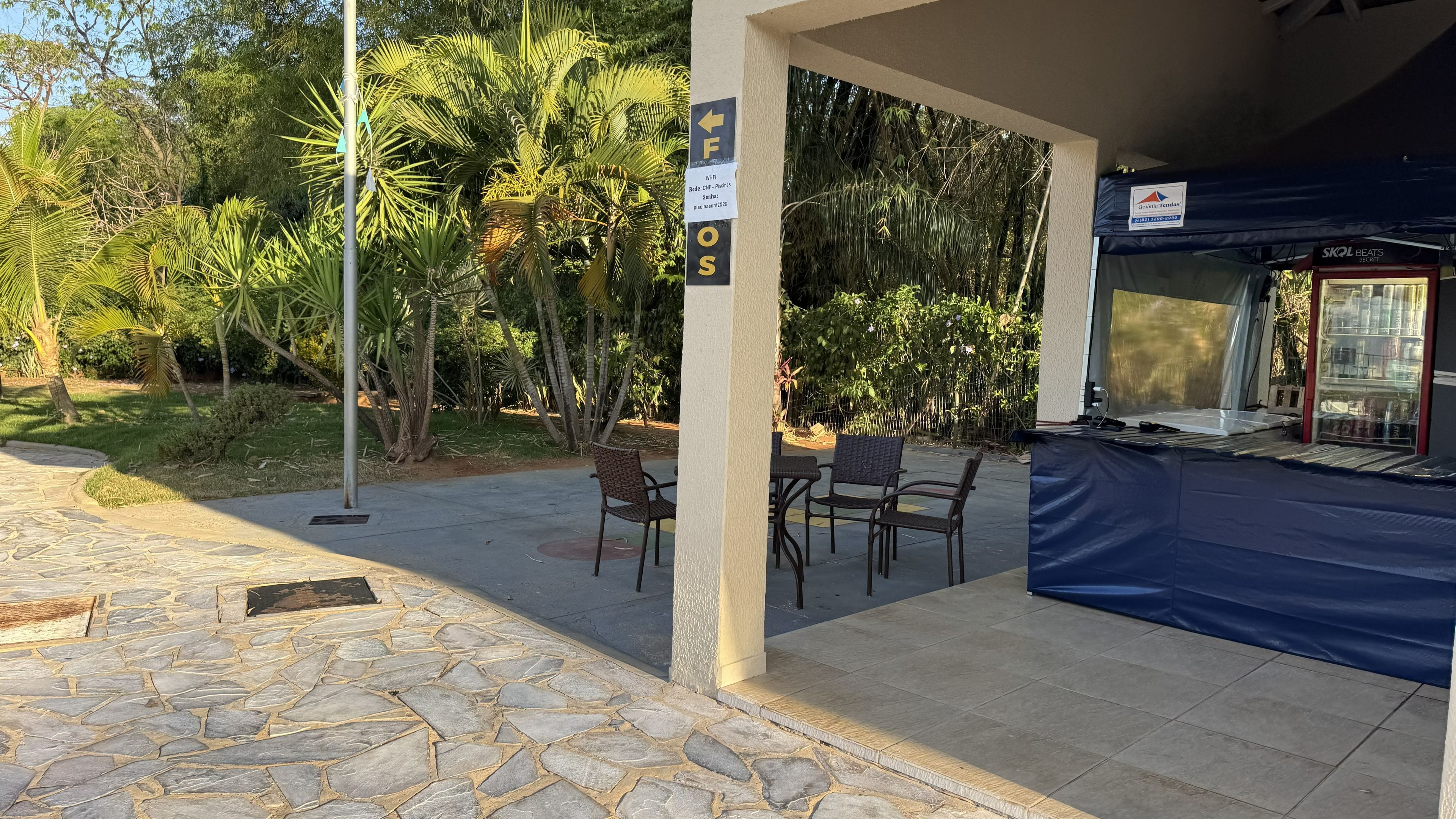 Uma área de estar externa com piso de pedra, uma seção coberta por uma lona azul e uma placa indicando disponibilidade de Wi-Fi.
