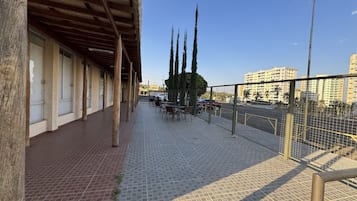 A covered outdoor seating area with tables and chairs, a tiled floor, and a view of a cityscape with buildings and a road.