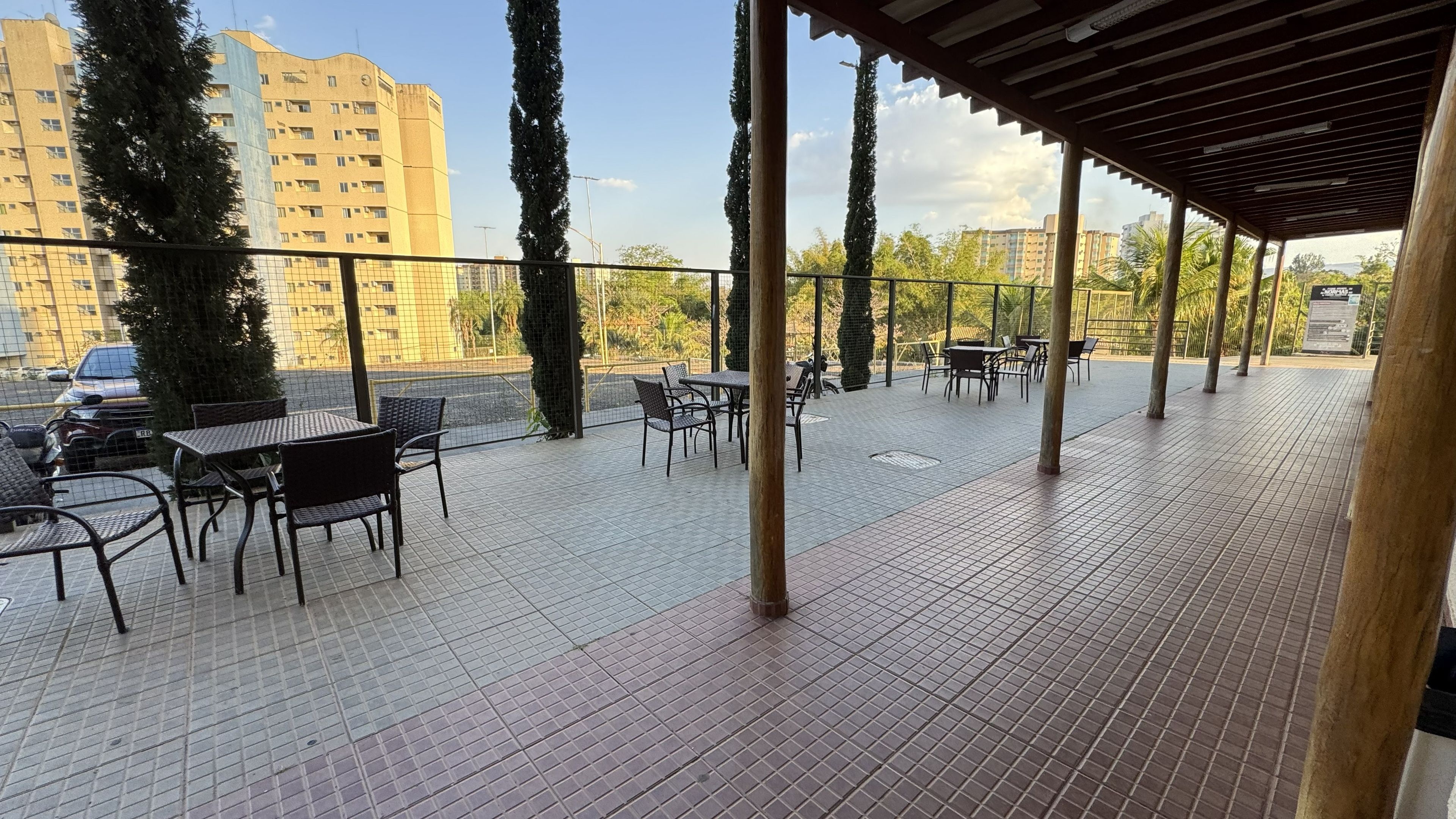 Un patio al aire libre con piso de baldosas, mesas y sillas de estructura metálica y altos árboles.