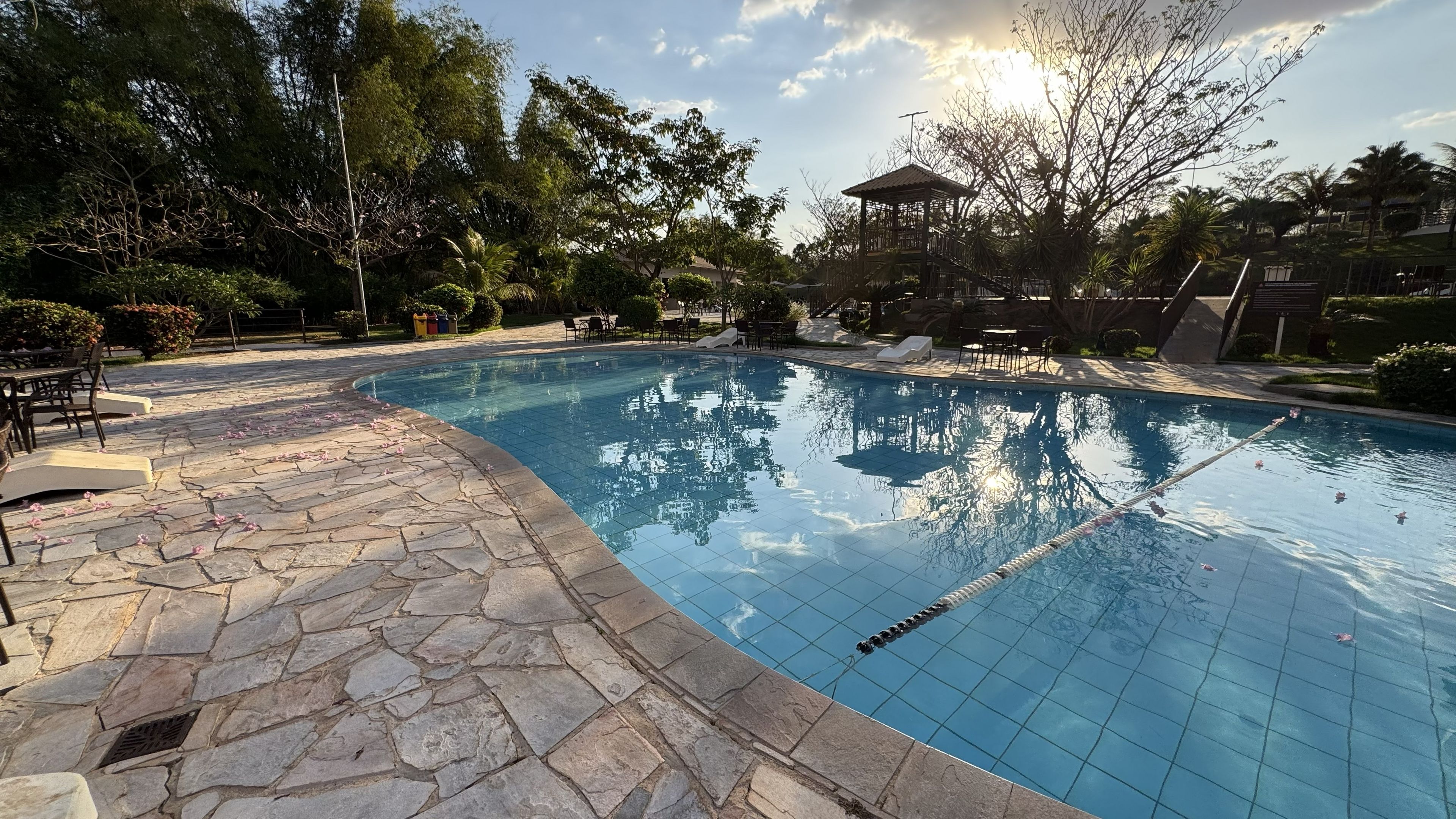Piscina com ladrilhos de pedra, rodeada por móveis de exterior e árvores.