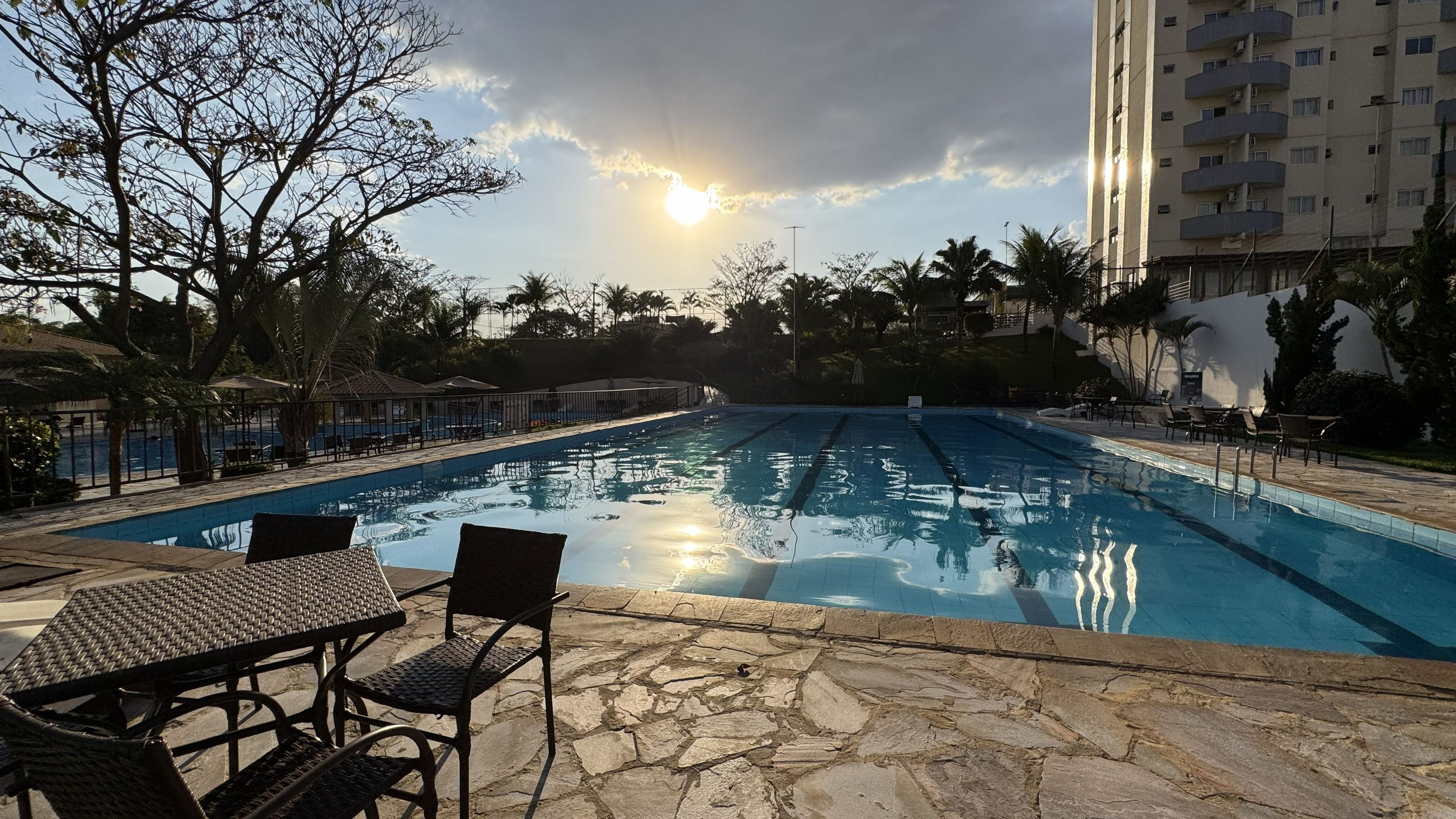 Una piscina con sillones de descanso y mesas, rodeada de palmeras y un edificio alto al fondo.