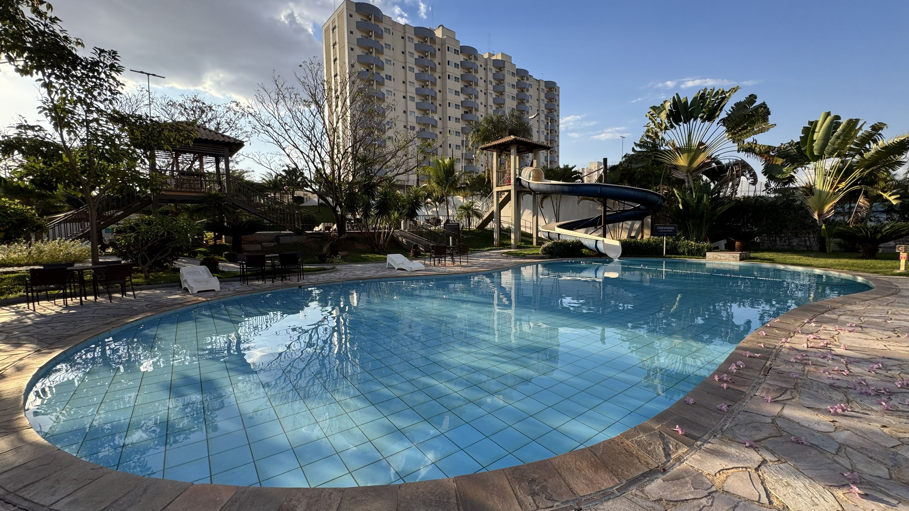 Una piscina con borde de azulejos, rodeada de sillones de descanso y un área de estar cubierta, con un edificio alto al fondo.