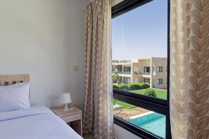 Ein Hotelzimmer mit Bett, Nachttisch mit Lampe, Fenster mit Vorhängen und Blick auf ein Gebäude und ein Schwimmbad.