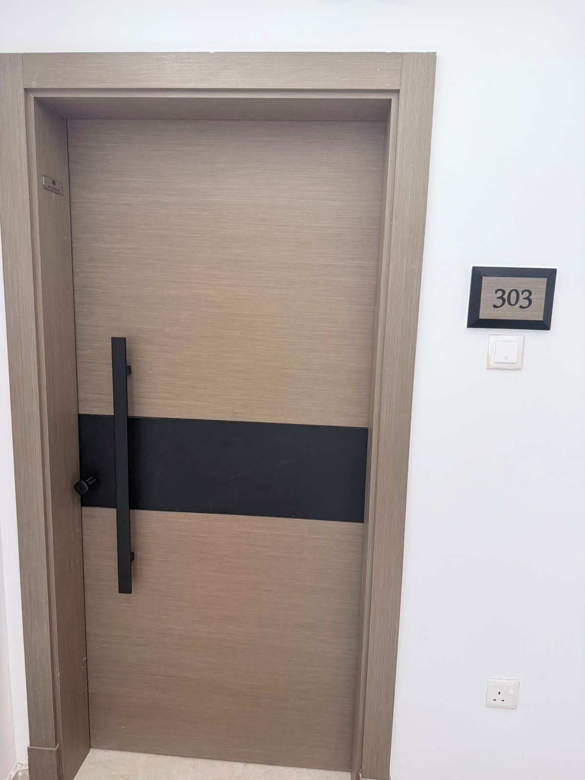 Une porte avec une bande horizontale noire, une plaque portant le numéro « 303 » et un interrupteur sur le mur.
