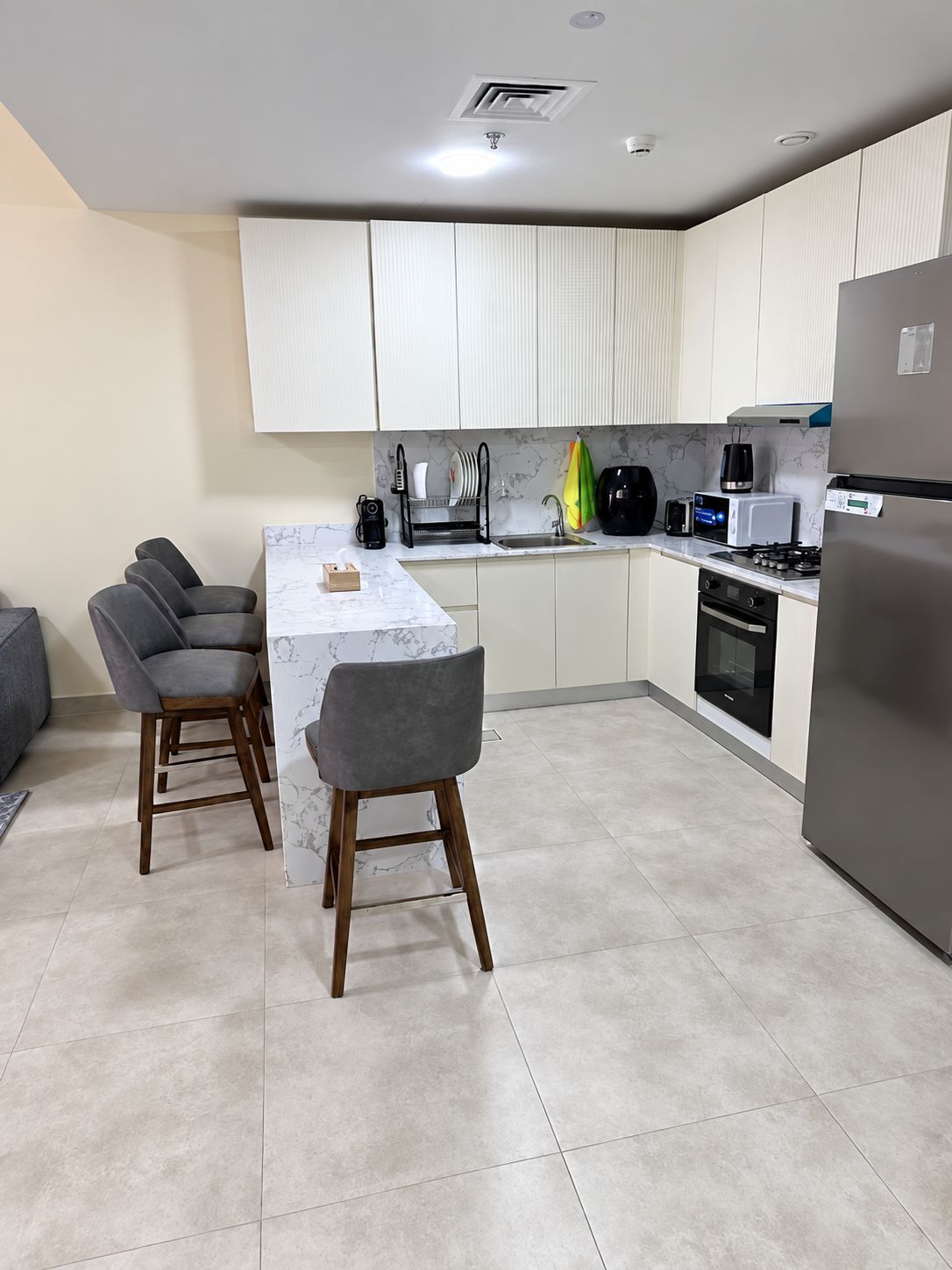 A modern kitchen with a marble countertop, two bar stools, a built-in oven, and a stainless steel refrigerator.