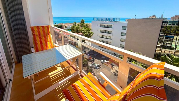 A balcony with a glass table and striped cushions, overlooking a cityscape and the sea. - Vrbo Property (Torremolinos)