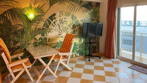 A room with a checkered floor, a table, two chairs, a TV, and a large tropical-themed mural. - Vrbo Property (Torremolinos)