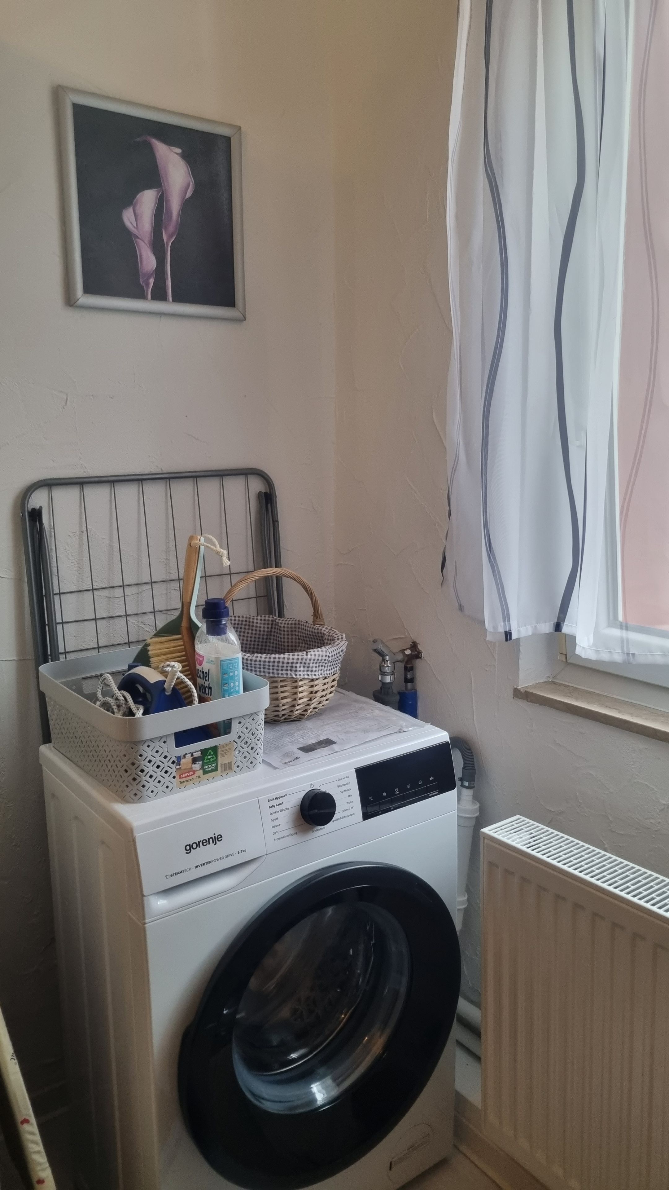 Ein Waschbereich mit Waschmaschine, Wäschekorb, einer Flasche und einem Bild an der Wand.
