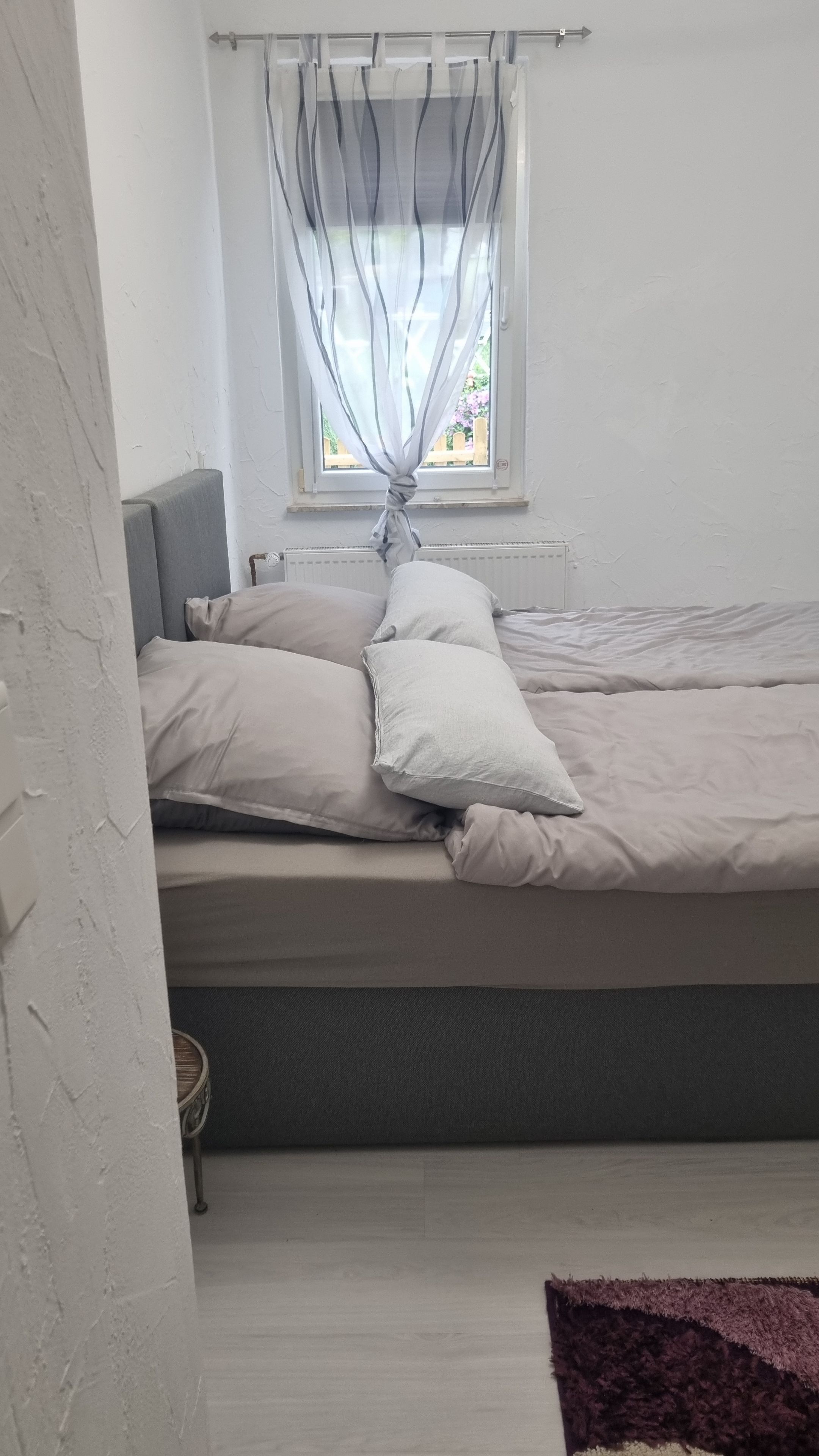 Ein Bett mit Kissen, ein Fenster mit Vorhängen und eine Heizung.