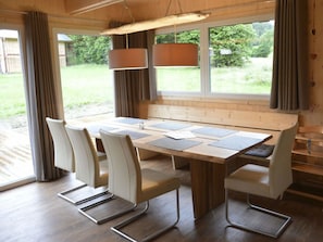 A dining area with a wooden table, white chairs, and a view of the outdoors through large windows. - One-bedroom Cabin (Sankt Georgen am Kreischberg)