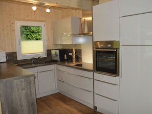 A modern kitchen with stainless steel appliances, wooden cabinets, and a window with blinds. - One-bedroom Cabin (Sankt Georgen am Kreischberg)