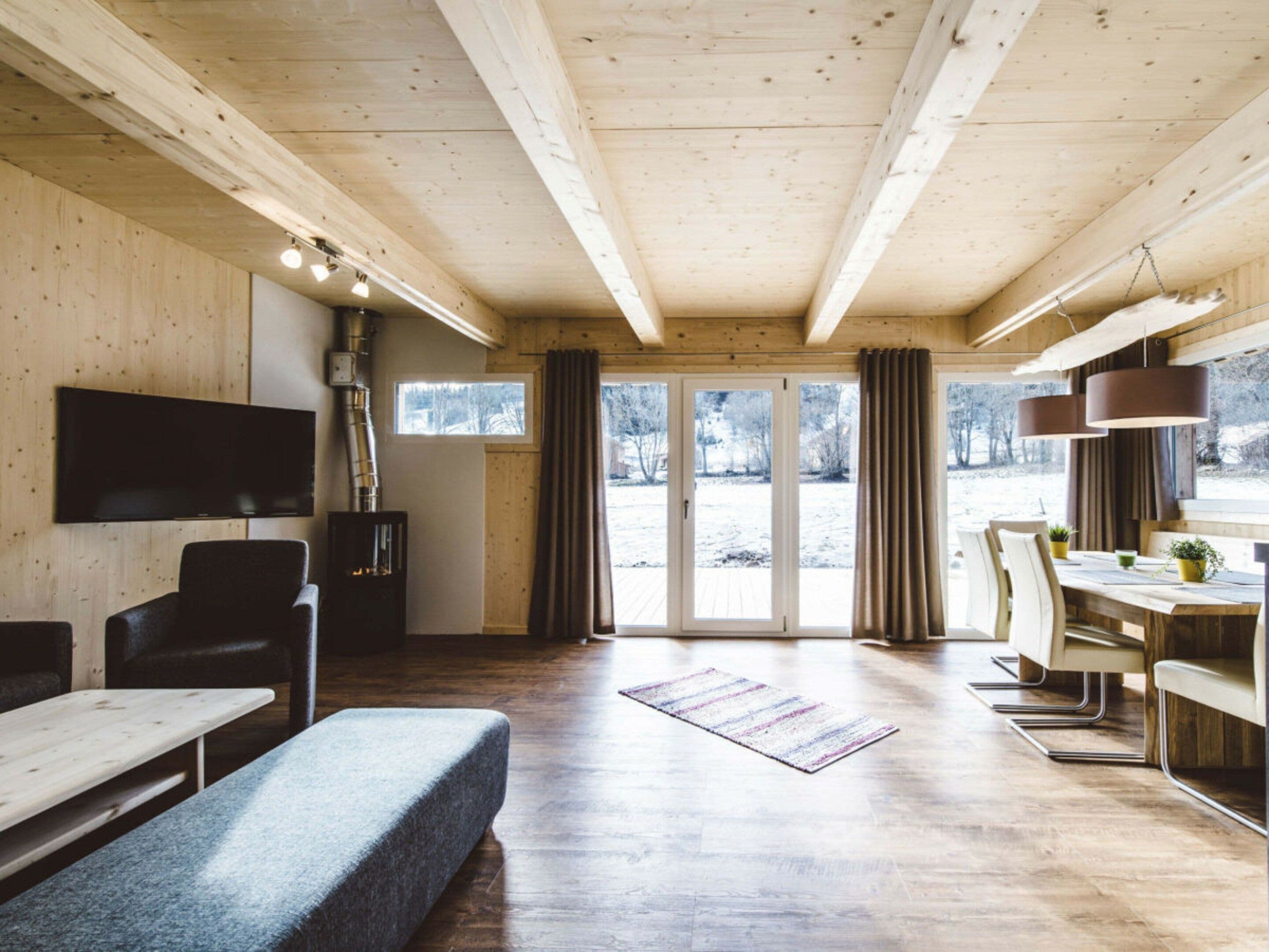 A modern interior with a wooden ceiling, a flat-screen TV, a fireplace, a dining area, and a view of a snowy landscape through large windows.