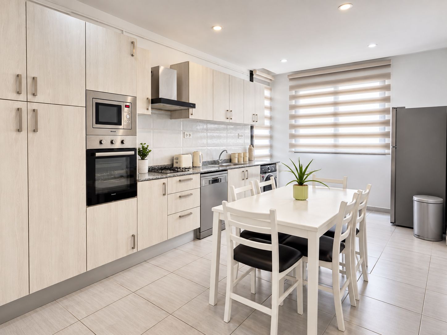 Una cocina moderna con armarios de madera clara, electrodomésticos de acero inoxidable, una mesa de comedor con sillas y una ventana con persianas.