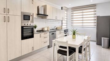 Una cocina moderna con armarios de madera clara, electrodomésticos de acero inoxidable, una mesa de comedor con sillas y una ventana con persianas.