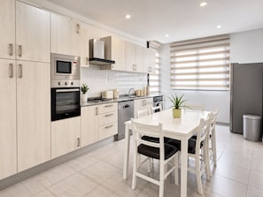 Una cocina moderna con armarios de madera clara, electrodomésticos de acero inoxidable, una mesa de comedor con sillas y una ventana con persianas.