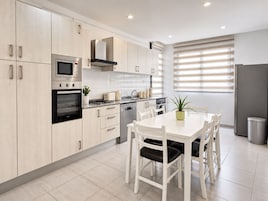 Una cocina moderna con armarios de madera clara, electrodomésticos de acero inoxidable, una mesa de comedor con sillas y una ventana con persianas.