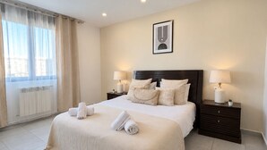 A hotel room with a large bed, two bedside tables with lamps, a radiator, and a window with curtains. - Spacieux 3 chambres Boulevard des Lions (Bir El Djir)