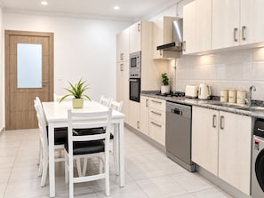 Una cocina moderna con muebles blancos, una mesa de comedor con sillas, horno empotrado, microondas, lavavajillas y cafetera.