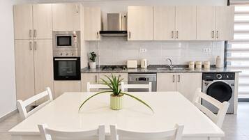 A modern kitchen with light wood cabinets, stainless steel appliances, a dining table with chairs, and a potted plant.