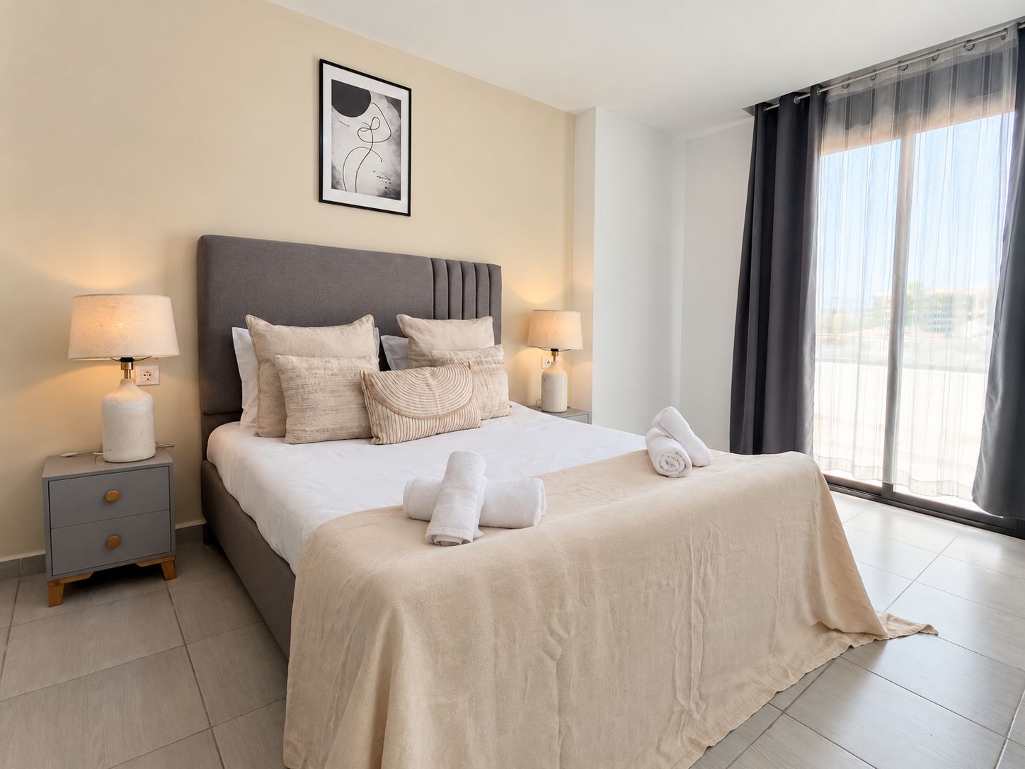 Habitación de hotel con cama, dos mesitas de noche, una lámpara de noche, ventana con cortinas y un cuadro en la pared.