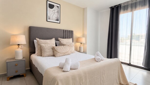 A hotel room with a bed, two bedside tables, a nightstand, a window with curtains, and a framed picture on the wall. - Spacieux 3 chambres Boulevard des Lions (Bir El Djir)