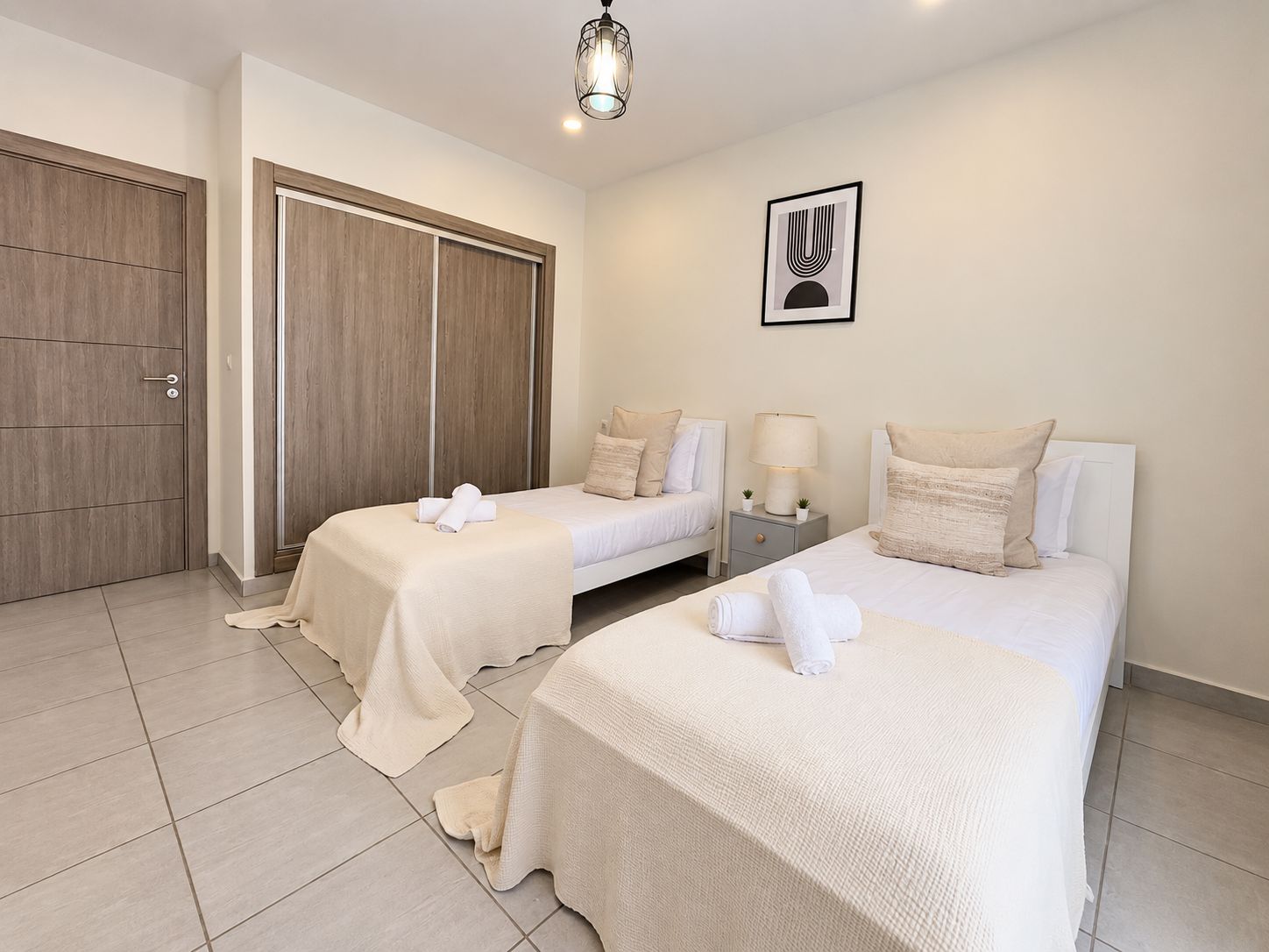 Habitación de hotel con dos camas, un armario, una mesita de noche con lámpara y un cuadro en la pared.