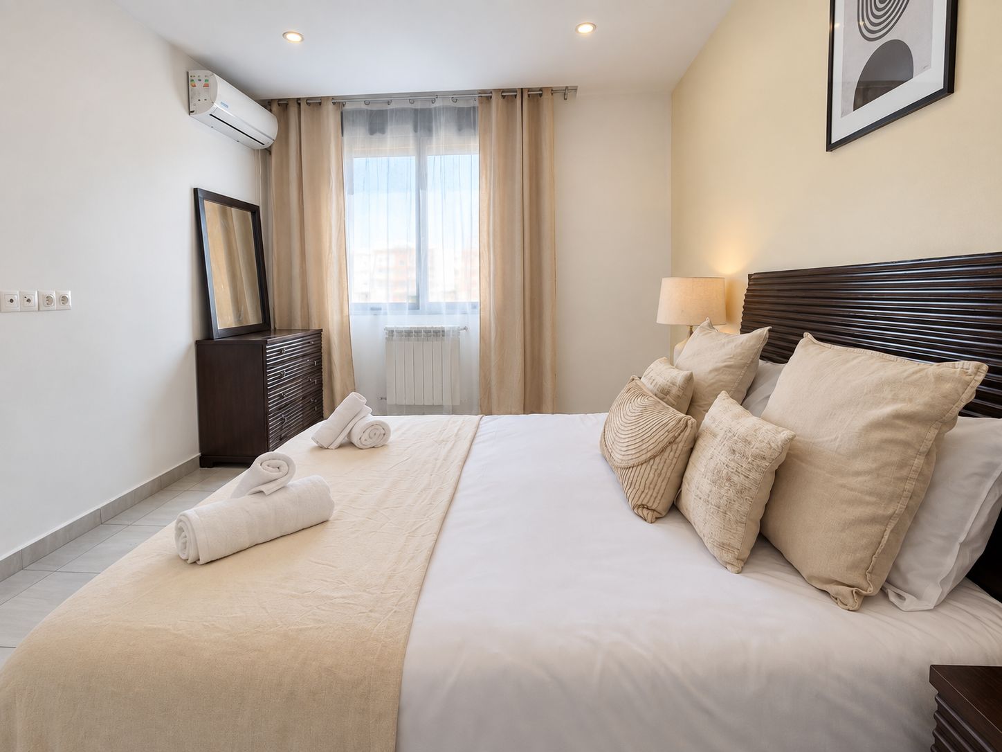 Una habitación de hotel con una cama, una lámpara de noche, una ventana con cortinas, un radiador y un equipo de aire acondicionado.