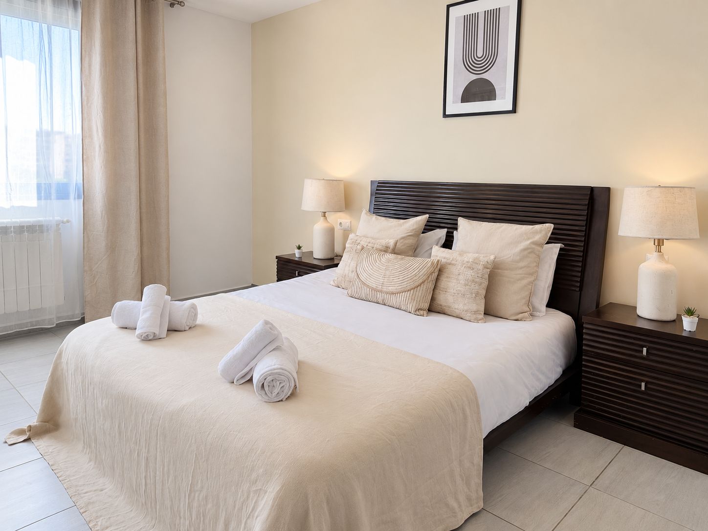 A hotel room with a large bed, two bedside tables, a wall-mounted lamp, and a framed artwork above the bed.