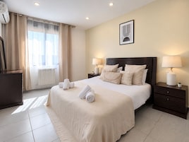 Una habitación de hotel con cama, lámparas de noche, un radiador, una ventana con cortinas y un cuadro colgado en la pared.