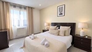 A hotel room with a large bed, two bedside tables with lamps, a radiator, a window with curtains, and a framed picture on the wall. - Spacieux 3 chambres Boulevard des Lions (Bir El Djir)