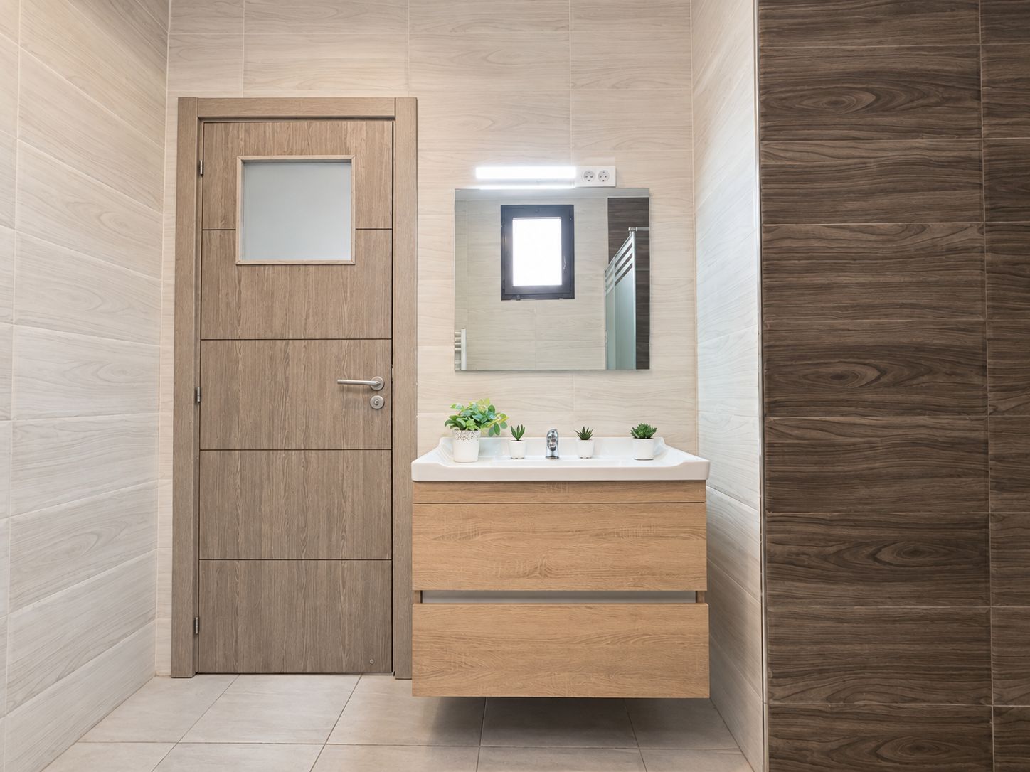 Un baño con puerta de madera, un espejo sobre un lavabo blanco y una ventana pequeña.