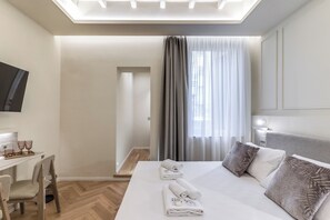 A hotel room with a bed, a desk, a chair, a TV, and a window with curtains. - Magenta Collection Sabbie (Rome)