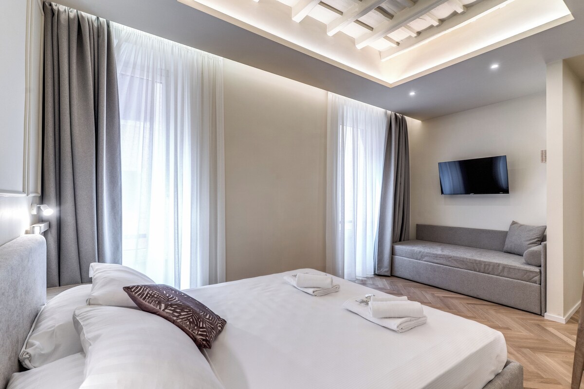A modern hotel room with a bed, a sofa, a flat-screen TV, and a ceiling light fixture.