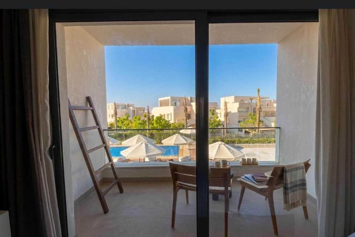 A balcony with a view of a pool and buildings, a wooden chair, a table, and a ladder.