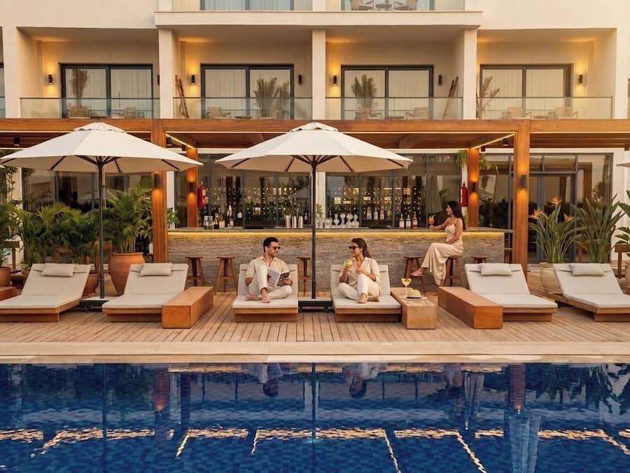 Uma área à beira da piscina com espreguiçadeiras, guarda-sóis, bar e piscina refletora.