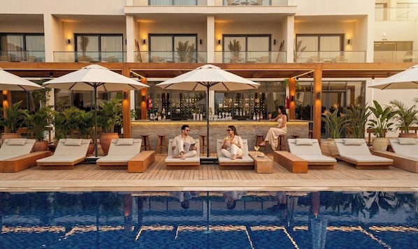 Un área junto a la piscina con sillones de descanso y sombrillas, un bar y una piscina reflectante.
