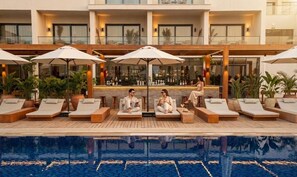 Área da piscina com espreguiçadeiras e guarda-sóis, um bar e reflexos na água.