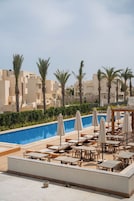 Zona de piscina en la azotea con sillones de descanso y sombrillas, rodeada de palmeras y edificios.