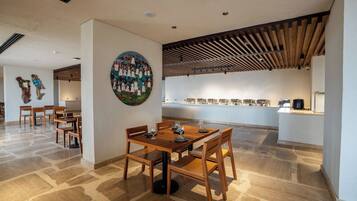 Un comedor con mesas y sillas de madera, un cuadro grande en la pared y una zona de cocina con una barra larga.