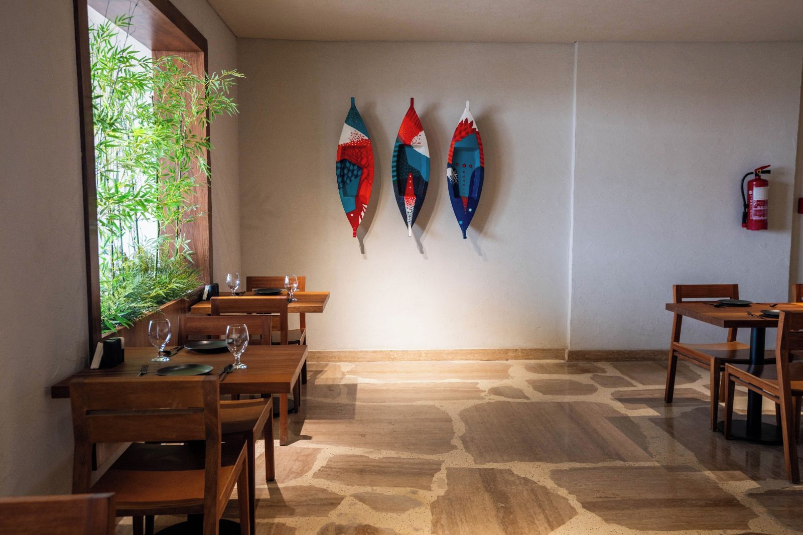 Un espace repas avec des tables et des chaises en bois, un extincteur fixé au mur et trois planches de surf colorées accrochées au mur.