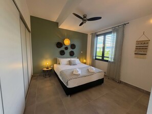 A bedroom with a bed, a ceiling fan, a window with curtains, and wall decor. - Tropical Elegance - 2-Bedroom Apartment with Terrace, Walking Distance to the Beach in L'Ermitage (Saint gilles les bains)