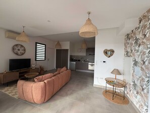 A modern living room with a sofa, television, and kitchen in the background. - Tropical Elegance - 2-Bedroom Apartment with Terrace, Walking Distance to the Beach in L'Ermitage (Saint gilles les bains)