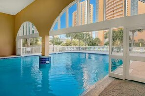 A swimming pool with clear blue water, surrounded by a white railing, and large windows offering views of tall buildings and palm trees. - Celadon 1505 - Turquoise Oasis (Panama City Beach)