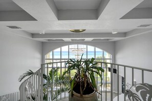 A spacious interior with a large arched window overlooking a beach, a central potted plant, and a white railing. - Celadon 1505 - Turquoise Oasis (Panama City Beach)