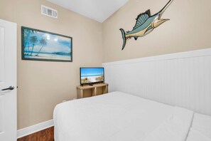 A bedroom with a bed, a television on a wooden stand, a painting of a beach scene, and a wall-mounted sculpture of a fish. - Celadon 1505 - Turquoise Oasis (Panama City Beach)